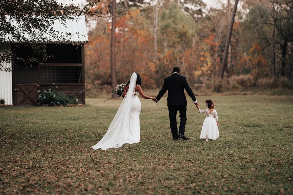 Bride and groom walking across a field holding hands with their flower girl. 