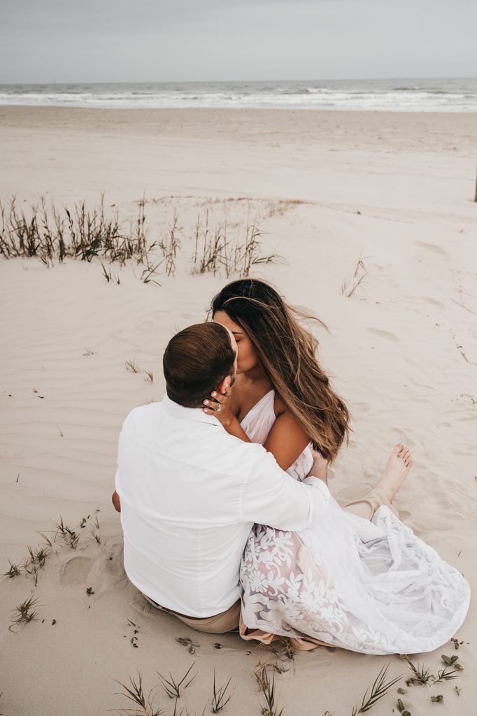 Woman in long dress straddling man on the sand and kissing him for a photo shoot. 