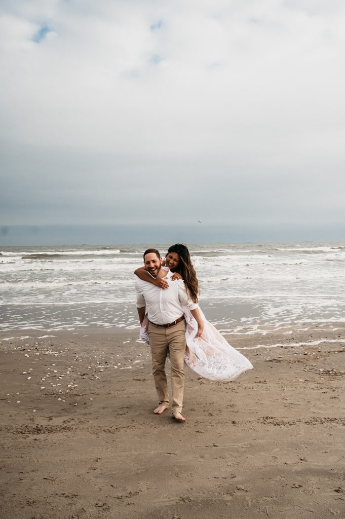 Woman riding on mans back across the beach for their engagement photos with Galveston elopement photographer, Brittany Emmanuel. 