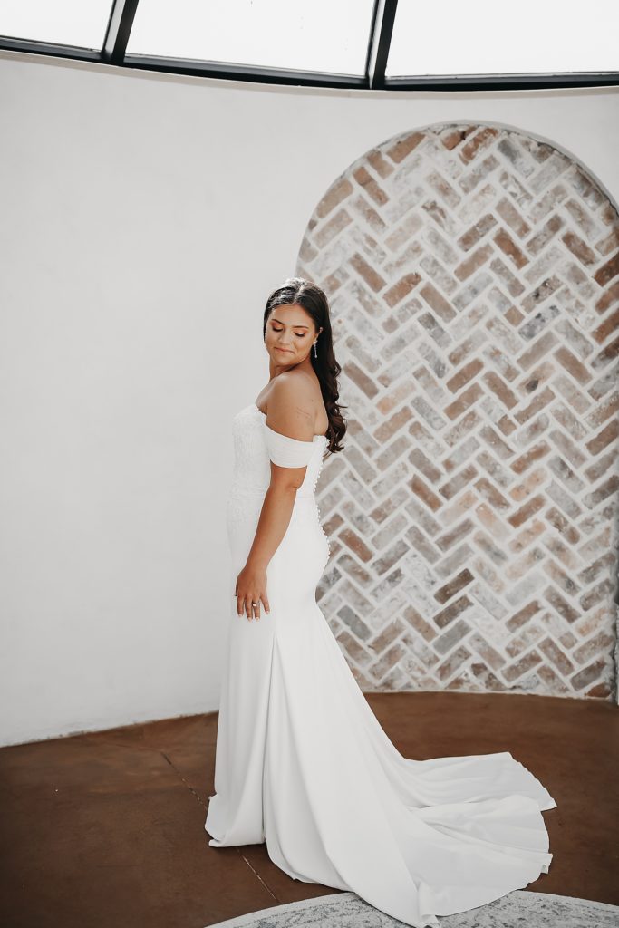 Bride in an off the shoulder white dress with a train in front of a exposed brick arch. 