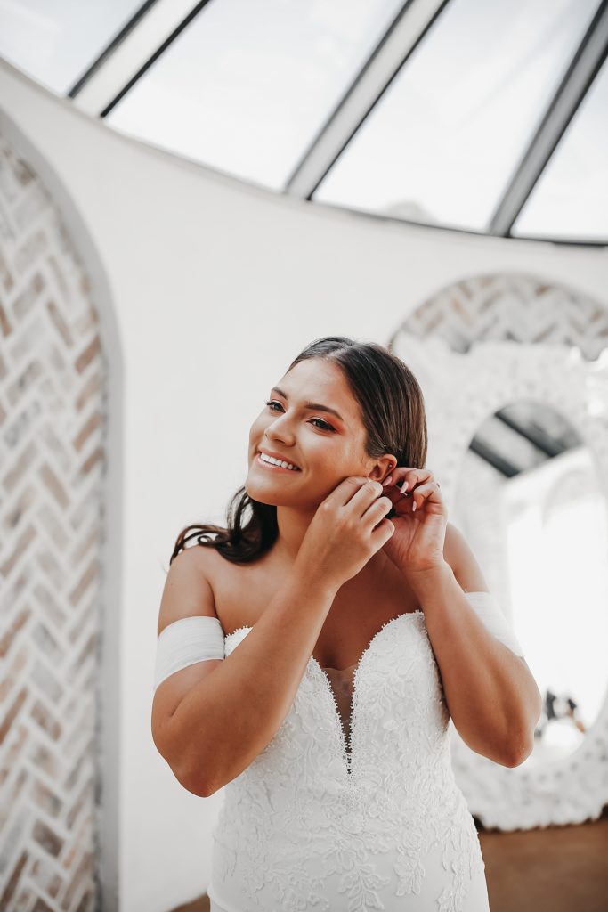 Bride putting on her earrings in a an atrium by Houston Wedding Photographer, Brittany Emanuel. 