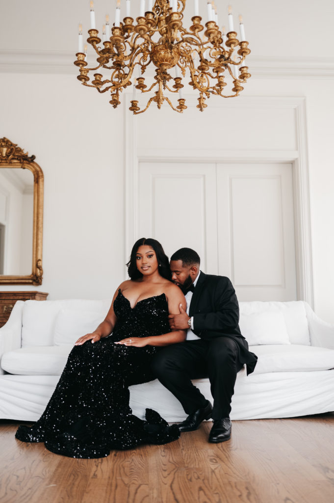 Engaged couple sitting on a white couch under a chandelier at The Creative Chateau
